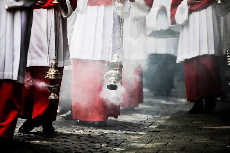 Meninos com vestes vermelhas e brancas, balançando incensários de metal.